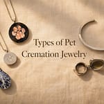Five types of pet cremation jewelry arranged on cream linen, including a glass-infused pendant, resin paw print disc, stainless steel paw chamber pendant, locket, and cremation bracelet